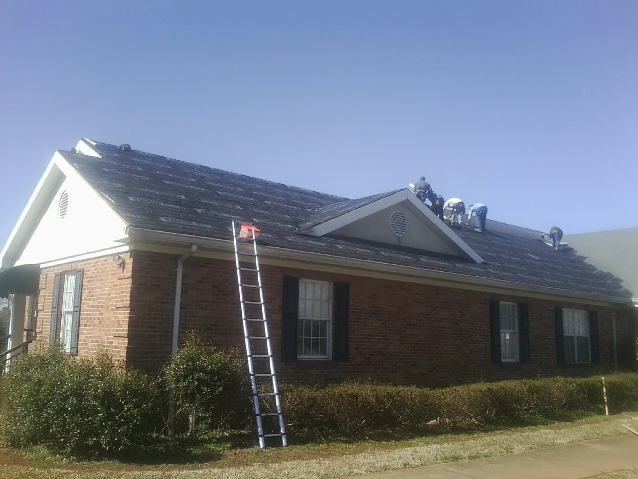 Crew tearing shingles off a brick ranch roof with a ladder set — Emergency Roof Repair work by our crew in Pollock
