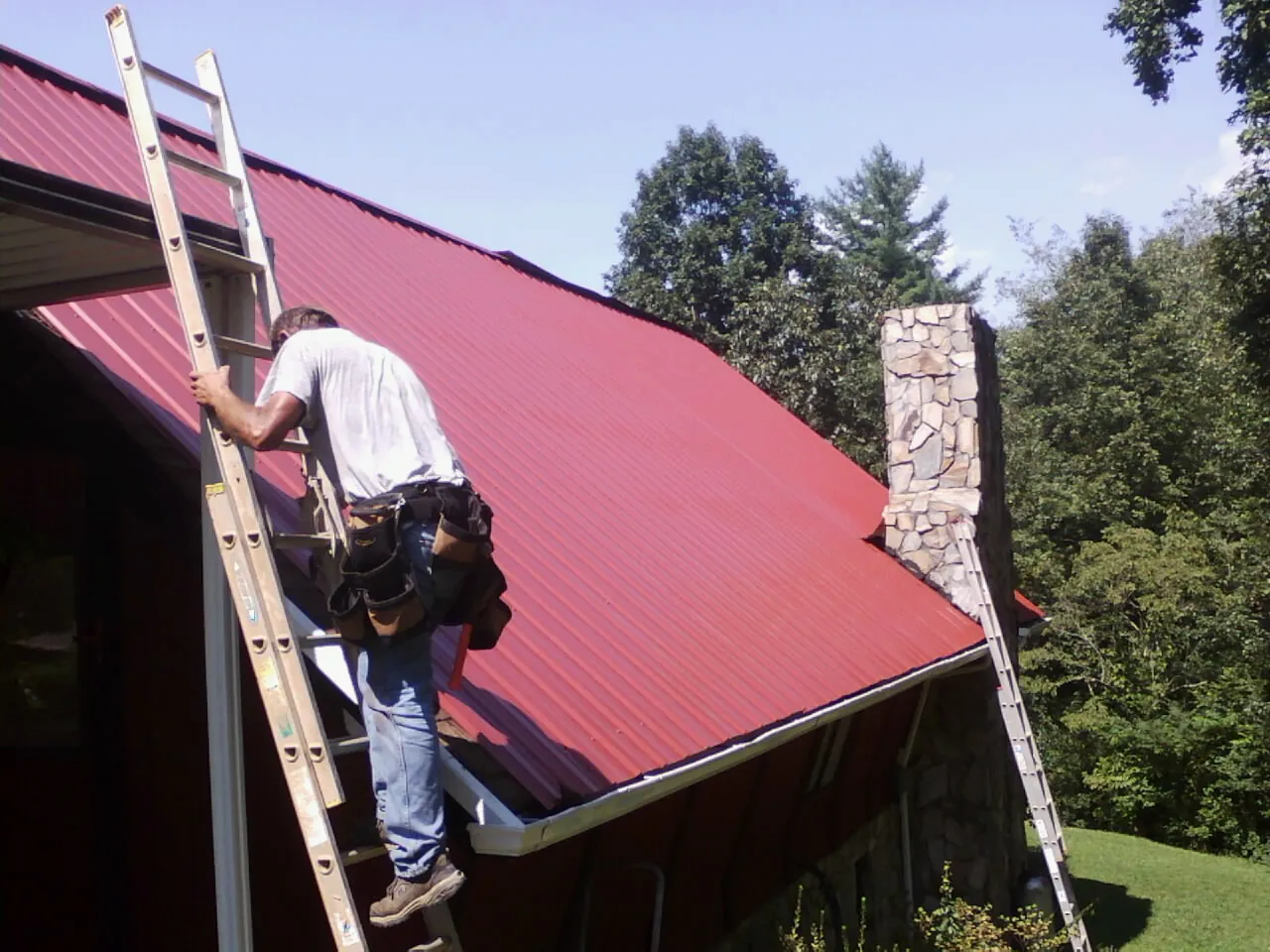 Roofer on a ladder beside a red metal roof and stone chimney — Soffit & Fascia Repair by Roofing Brothers in Pollock